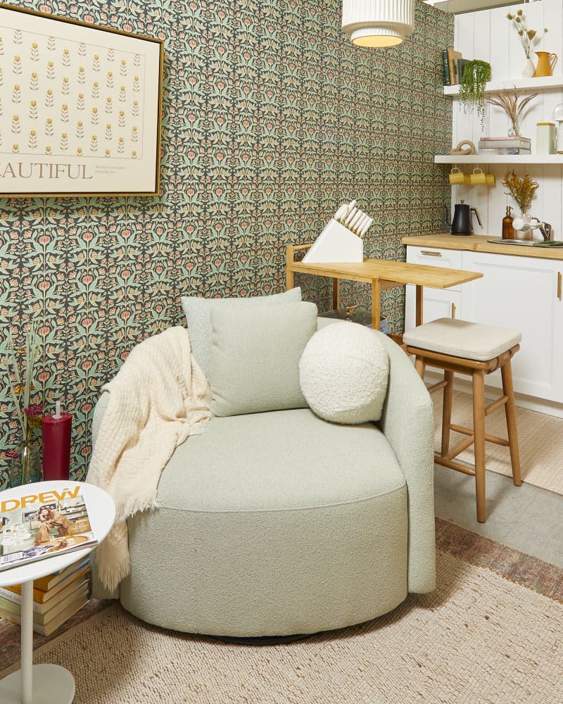 Head on view of a light green swivel chair with a white throw blanket and a white spherical pillow.  The wall behind it has a sage and light pink floral wallpaper.