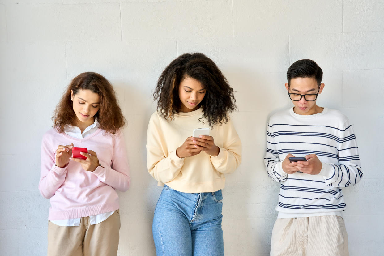 Young teenage cool multiethnic group of gen z high school college students standing on white wall background holding mobile cell phones looking at smartphones using apps. Social media concept