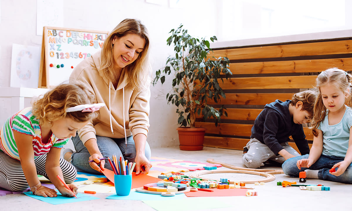 A teacher sits on the floor with two young students, who are coloring and playing with small toys.