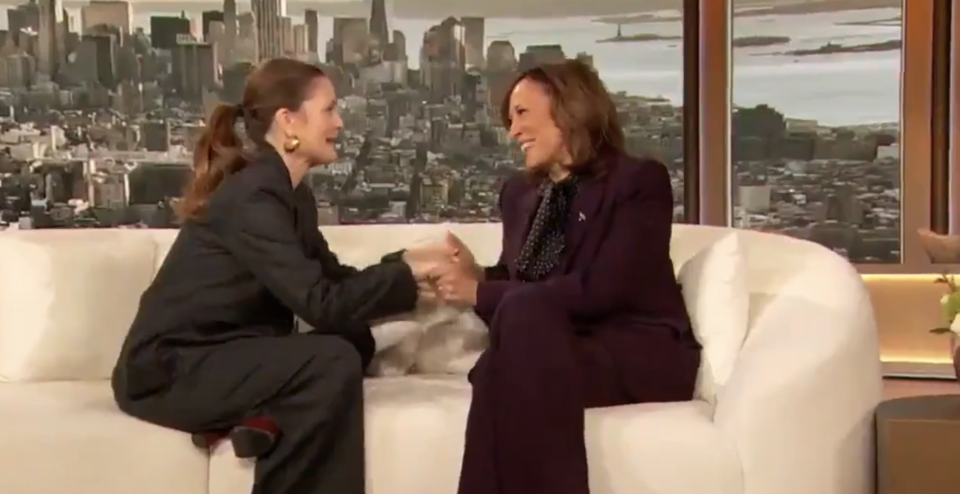Two women shaking hands on a talk show set, one seated on a white couch, both smiling