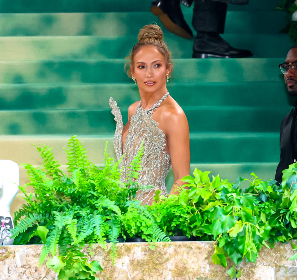 Jennifer Lopez in a sparkling gown with a deep neckline, hair in a bun, standing on steps with greenery