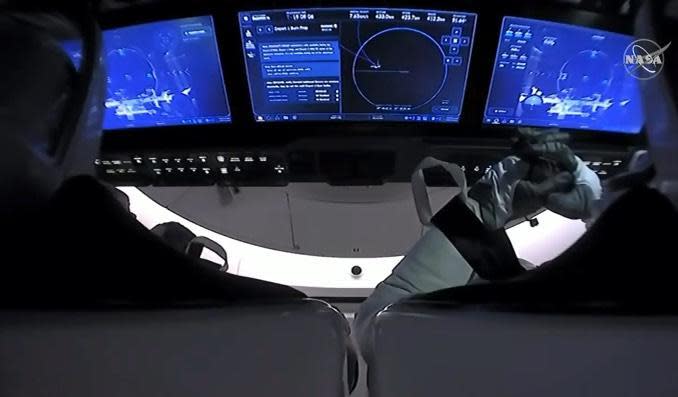 An over-the-shoulder view of the Crew Dragon's three touchscreen displays, used to command the spacecraft and monitor vehicle performance, showing external camera views of the receding space station. Hurley is strapped in to the left with Behnken to the right. / Credit: NASA/SpaceX