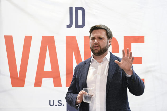 U.S. Senate candidate J.D. Vance, holding a plastic cup of water, speaks with a poster showing his name in the background.