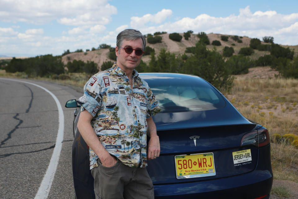 Howard Coe poses in front of his wife's Tesla sedan on Tuesday, Sept. 14, 2021, on a road near Nambé Pueblo outside of Santa Fe, N.M. Coe welcomed the opening of a new Tesla store and repair shop where he can pickup an SUV he's buying. Last year, he and his wife had to pick up her car in Denver, Colo., about five hours away, because of New Mexico laws that bar companies like Tesla from selling cars directly to customers. (AP Photo/Cedar Attanasio)