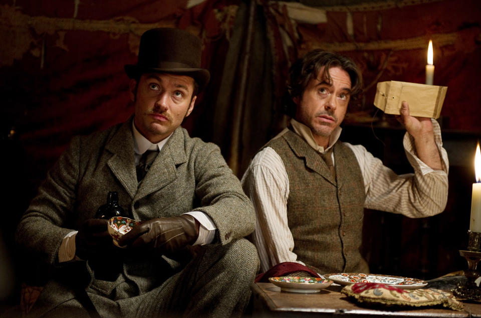 Jude Law and Robert Downey Jr. dressed in vintage attire, sitting indoors with dim lighting; Downey Jr. holds a document near a candle