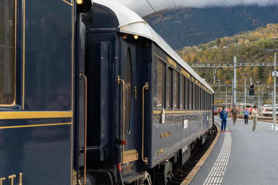 A navy blue train with gold trimmings stopped at a platform with mountains in the background