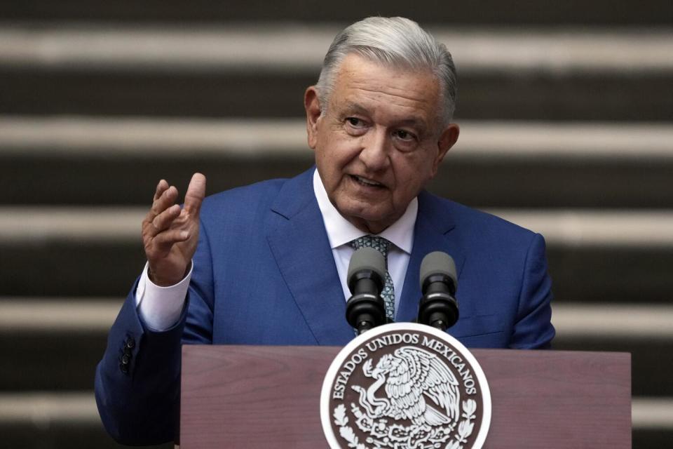 A man with gray hair, in a blue suit, gestures with one hand while speaking at a lectern with a government seal