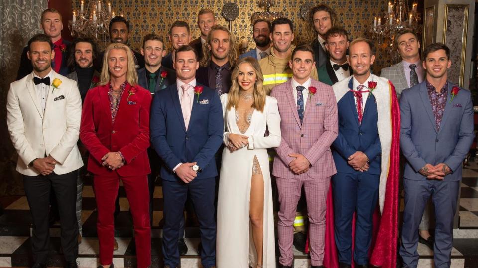 Bachelorette star Angie Kent wearing a white dress surrounded by male contestants in various different coloured suits