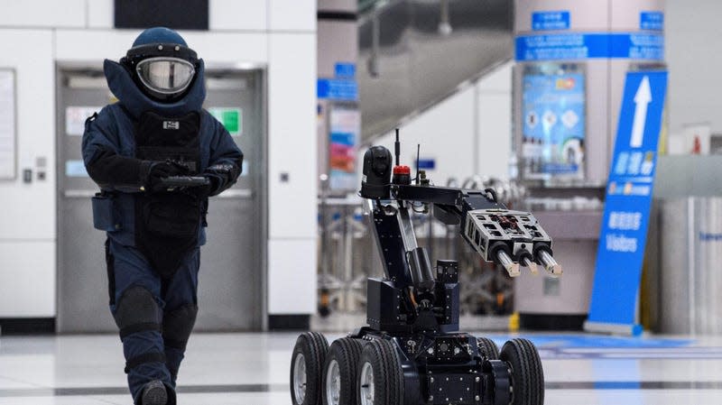 A police officer wearing full clear is seen with a remote control. A bomb disposal robot moves in front of them.