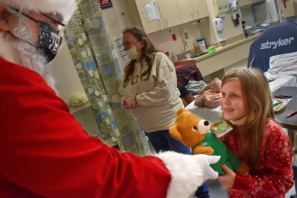 Santa hands a teddy bear to a delighted child in the hospital on Christmas Eve.