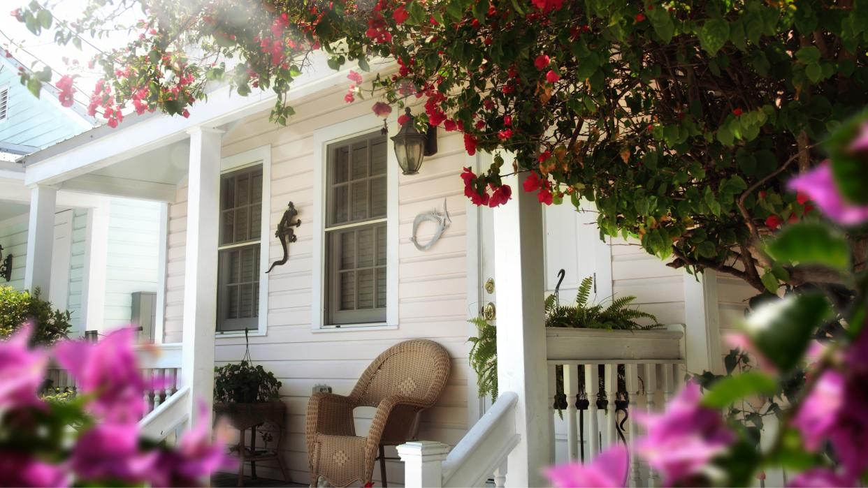  Summer shot of white wooden home with front porch wicker furniture and viewed through a frame of purple and red flowers and dapple sunlight. 