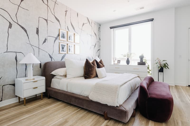 White bedroom with one gray and white patterned wallpaper wall, gray velvet bed, and other velvet accents