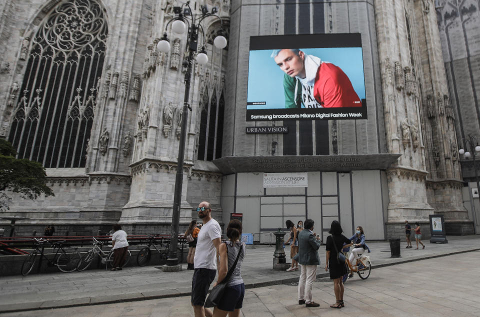 Pedestrians pass by a screen on the Duomo cathedral, showing a Moschino model during the Milan Digital Fashion Week, in Milan, Italy, Tuesday, July 14, 2020. Forty fashion houses are presenting previews of menswear looks for next spring and summer and pre-collections for women in digital formats, due to concerns generated by the COVID-19. (AP Photo/Luca Bruno)