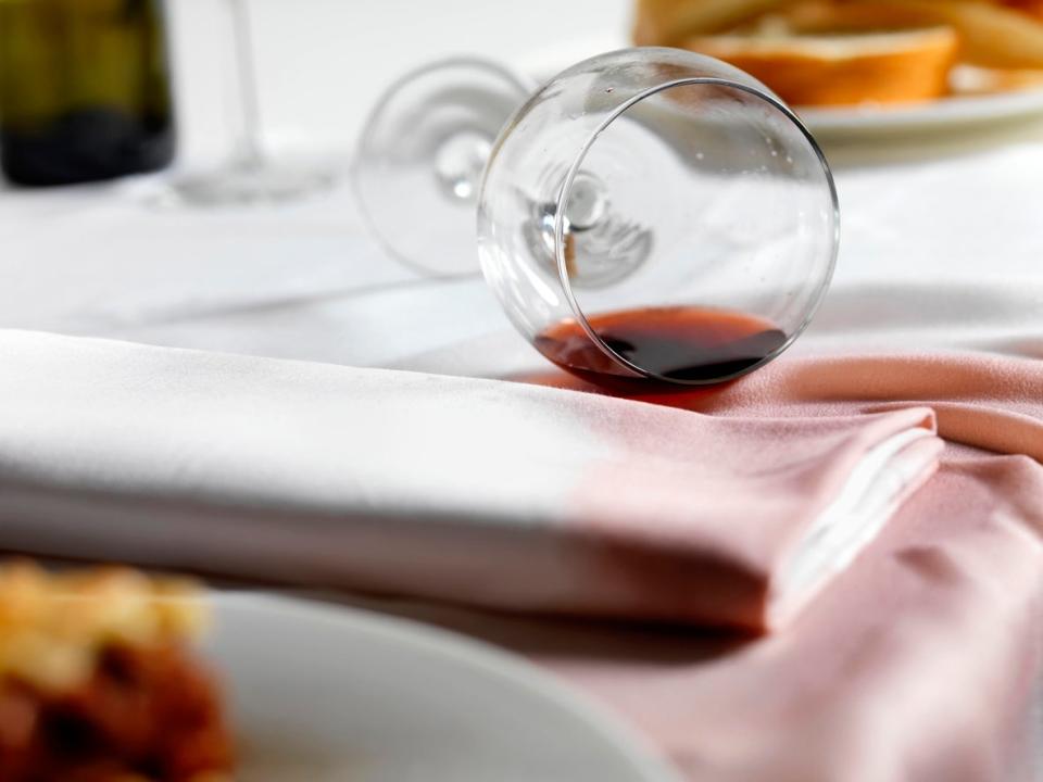 A glass of red wine spilled and staining a white tablecloth and napkin.