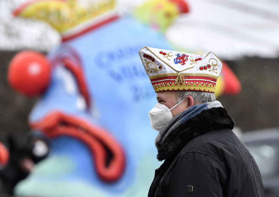 A reveller stands in front of political carnival floats to be shown in the streets of Duesseldorf, Germany, Monday, Feb. 15, 2021. Because of the coronavirus pandemic the traditional 'Rosenmontag' carnival parade are canceled but eight floats are pulled through the empty streets in Duesseldorf, where normally hundreds of thousands of people would celebrate the street carnival. (AP Photo/Martin Meissner)