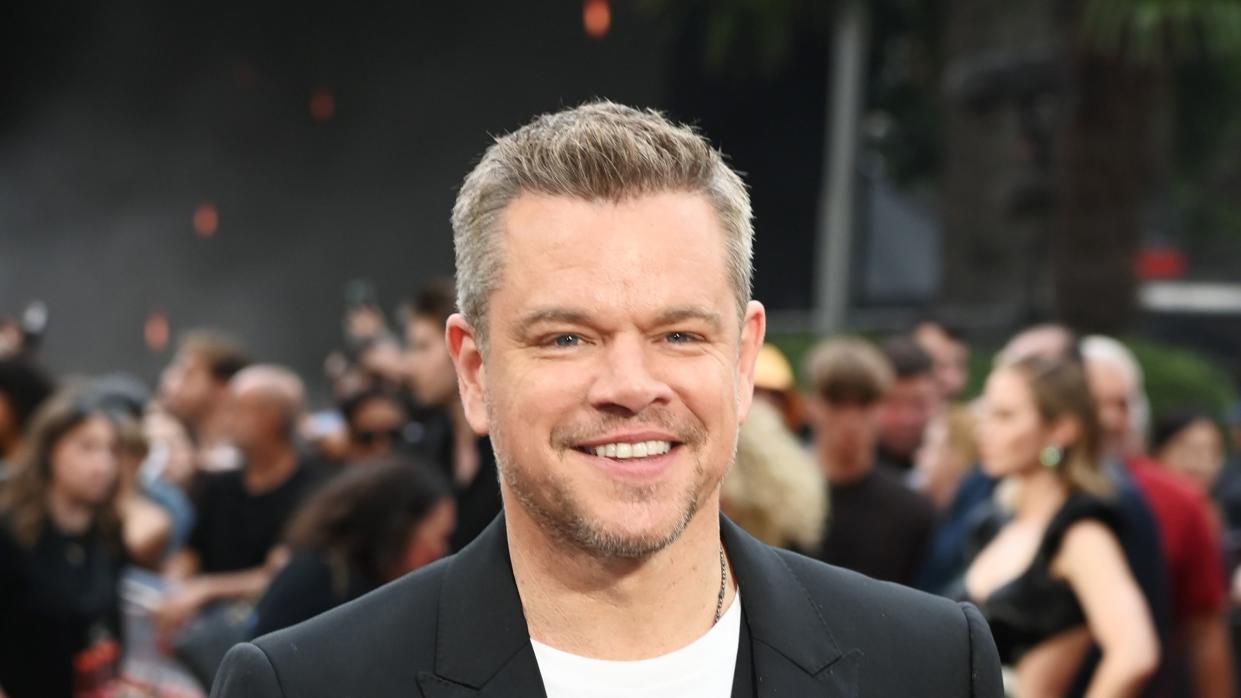 matt damon smiles at the camera, he wears a black suit jacket over a black vest and white shirt, behind him is a blurred out crowd of people