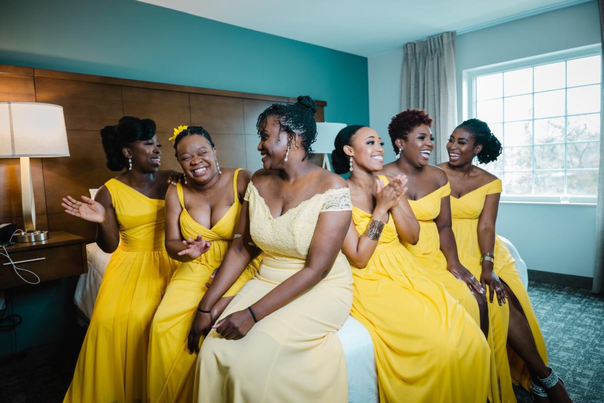 Black woman bride with bridesmaids in yellow dresses sitting on bed in hotel room