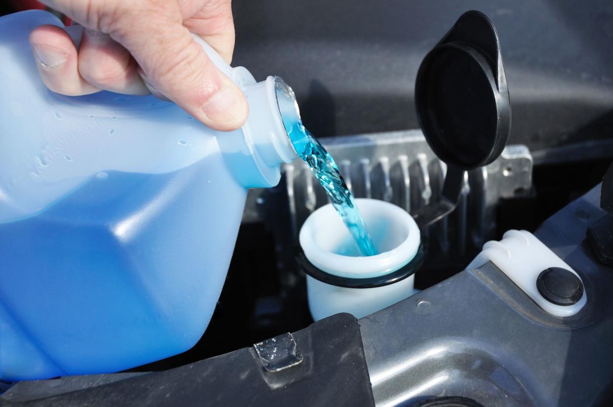 Closeup of a hand pouring a gallon jug of windshield washer fluid in the washer fluid reservoir under the hood of a car