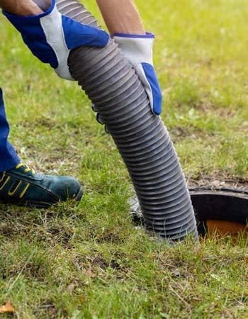 A person in blue and white closes inserts a tube down a sewer pipe in a yard. 