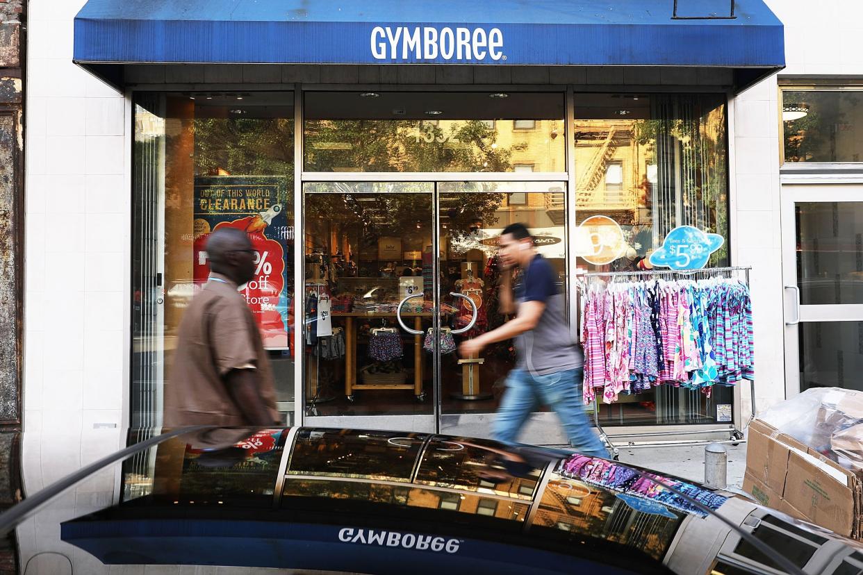 People briskly walk by front exterior of Gymboree store, Manhattan, New York City with a rack of children's clothes on the sidewalk with car in the foreground, it's windows reflecting the front sign