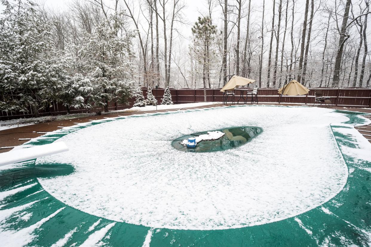 An in-ground swimming pool with a cover on it and water pump during a snowfall in winter in Indiana with evergreen and leafless trees in the background