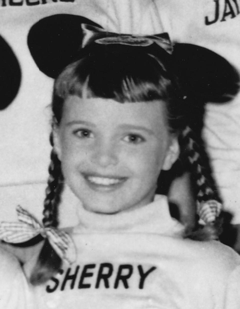 sherry alberoni smiles at the camera she wears a turtleneck with her first name embroidered on it and mickey mouse ears, her hair is in braided pigtails with bows at the ends