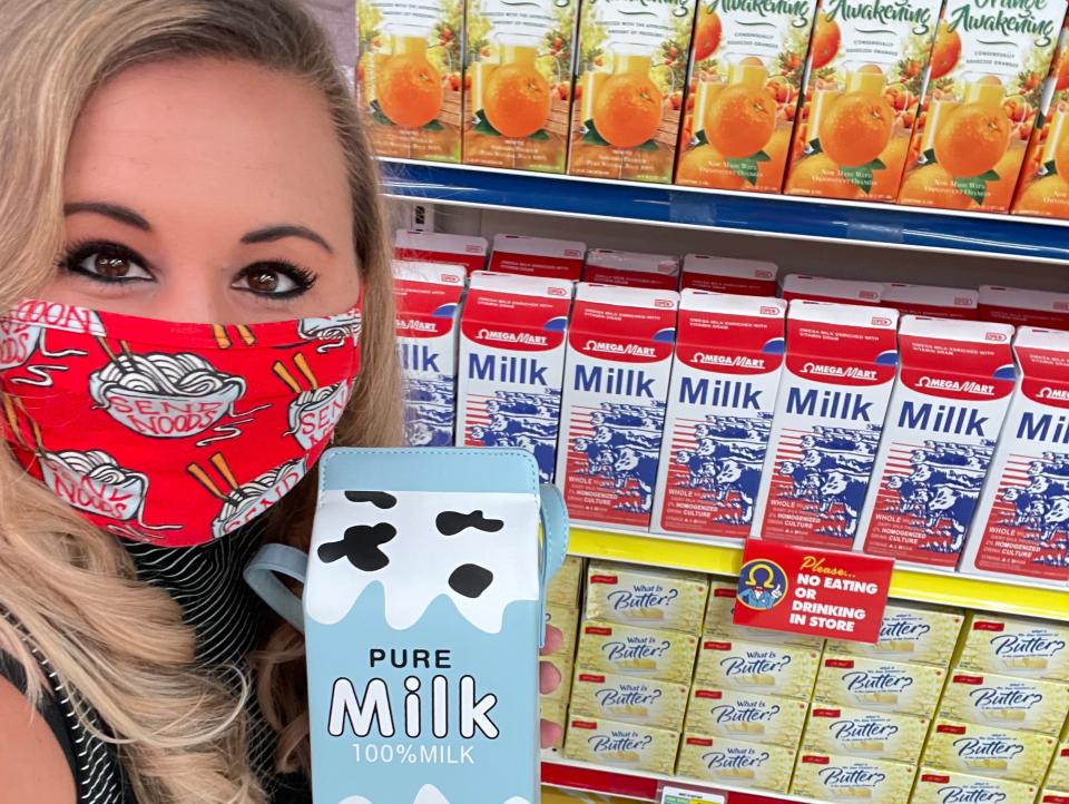 Carly Caramanna holding faux milk container and wearing face mask in front of aisle of faux groceries at omega mart