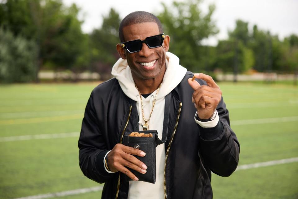 Deion Sanders in a white hoodie, black jacket, and sunglasses smiling on a football field while holding a black leather satchel of almonds.