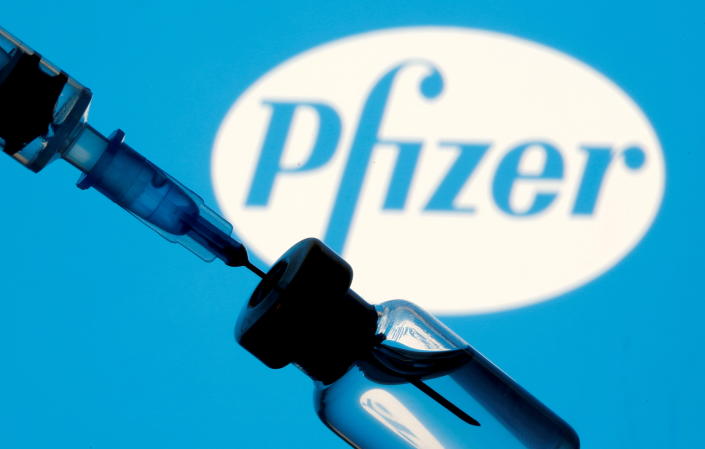 Un flacon et une seringue sont visibles devant un logo Pfizer affiché.