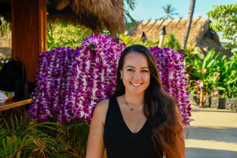 Niki Rickard stands smiling in front of a rack of purple flower leis