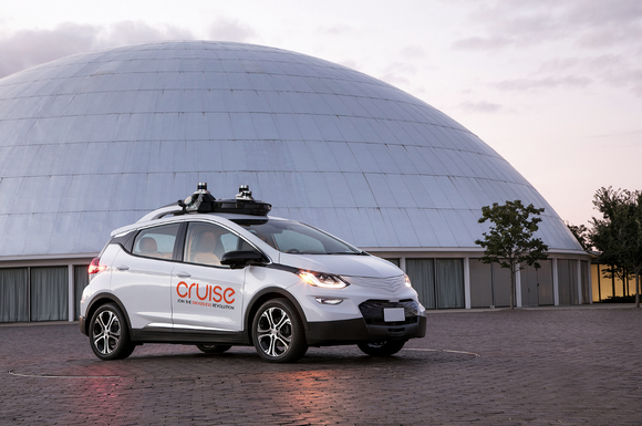 A white Chevrolet Bolt EV with Cruise logos and visible self-driving sensor hardware is parked outside GM's historic Design Dome in Warren, Michigan.