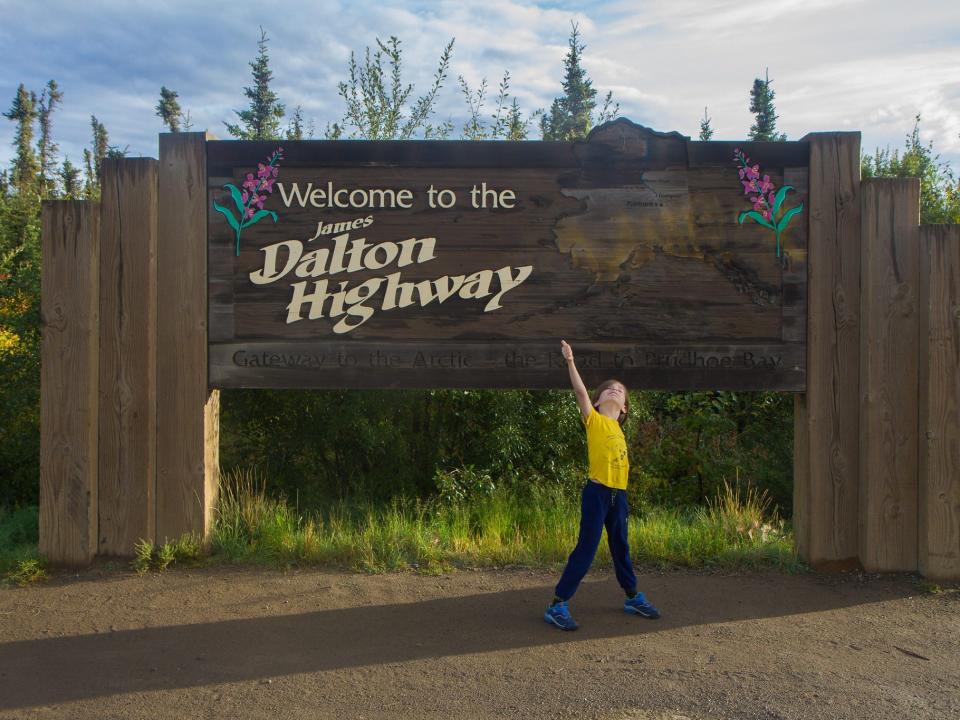 A boy giving a thumbs up in front of a sign for the Dalton Highway in Alaska.