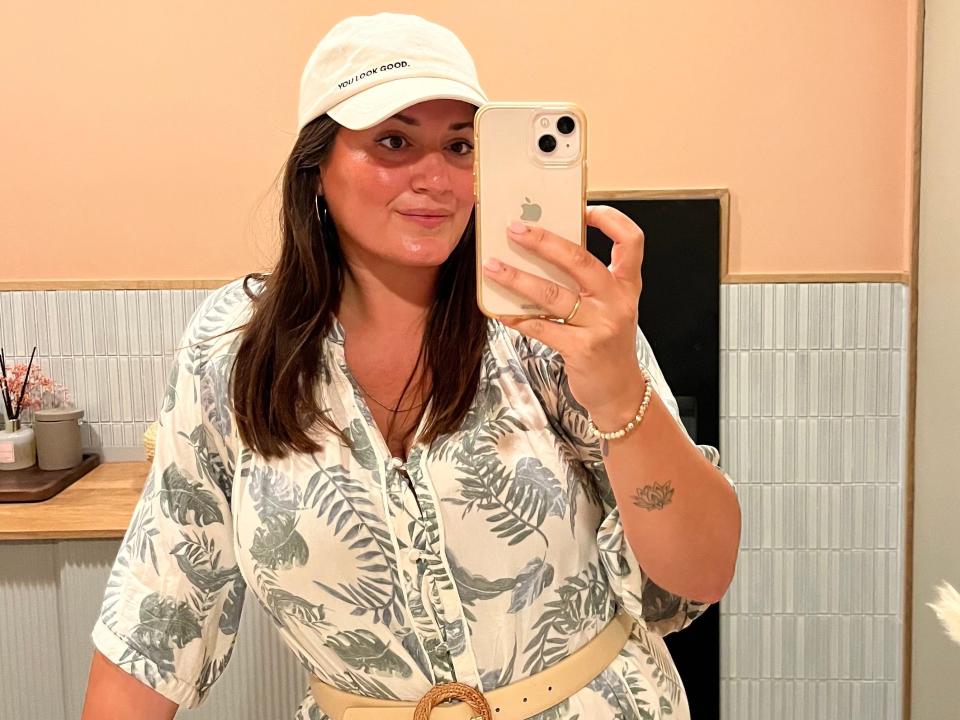 Lola Méndez takes a photo in the mirror with her iPhone. She has long dark brown hair, brown eyes, and wears a white baseball hat that says "you look good" in all caps on the front. She wears silver hoop earrings and a white top with a grey palm tree print.