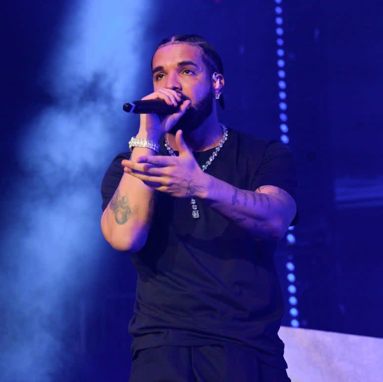 Drake performing on stage, holding a microphone. He is wearing a black t-shirt, a gold chain, and has tattoos on his arm