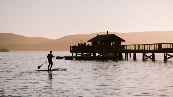 A paddle boarder at dusk on Tomales Bay, with the boat shack nearby