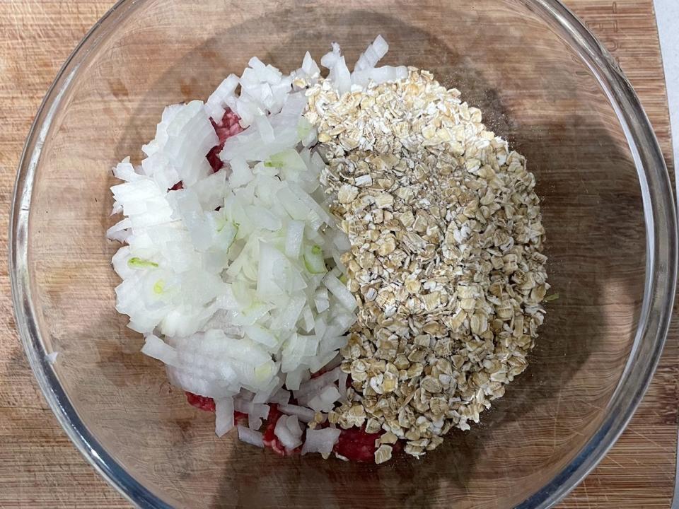 minced onions, ground meat, and oats in a glass mixing bowl on a wooden cutting board