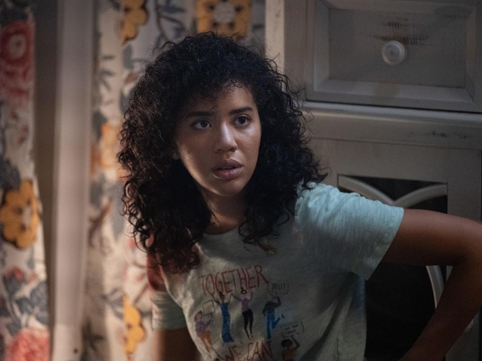 A still from Scream (2022) shows Jasmin Savoy Brown as Mindy Meeks-Martin sitting in a chair looking shocked