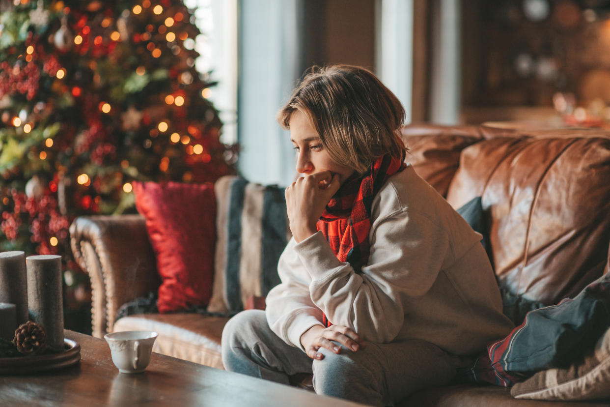 Young teen boy with long hair thoughtful look sad eyes negative mood angry mental health and crying at home. Stylish zoomer gen Z pensive on new year holidays with xmas tree bokeh lights garlands eve 25 december