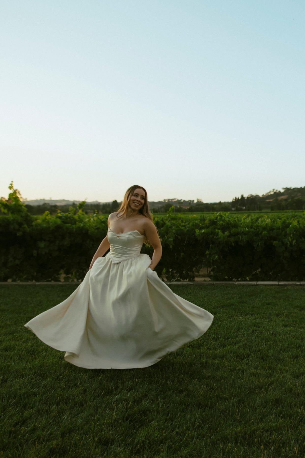 A bride stands in her wedding dress in a grassy field.