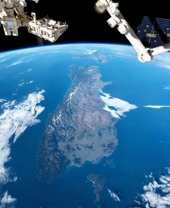 A satellite view of an island, likely New Zealand's South Island, surrounded by the ocean with visible mountain ranges and varied terrain. Space station parts appear at the edges