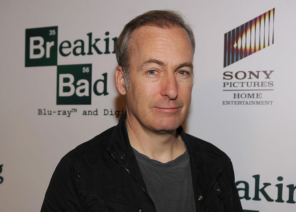 Bob Odenkirk stands in front of a "Breaking Bad" backdrop at an event, wearing a black jacket over a grey shirt