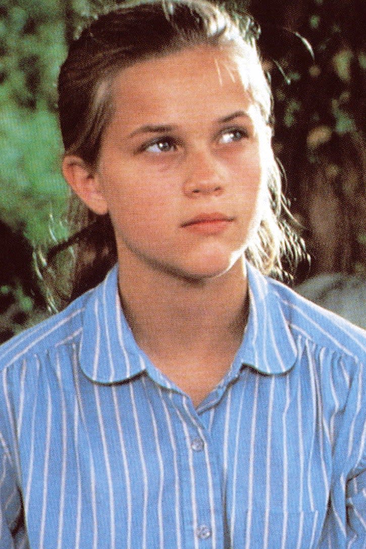 Reese Witherspoon in a striped shirt sits on the ground, looking up while holding another person who is resting their head on her lap in a grassy outdoor setting