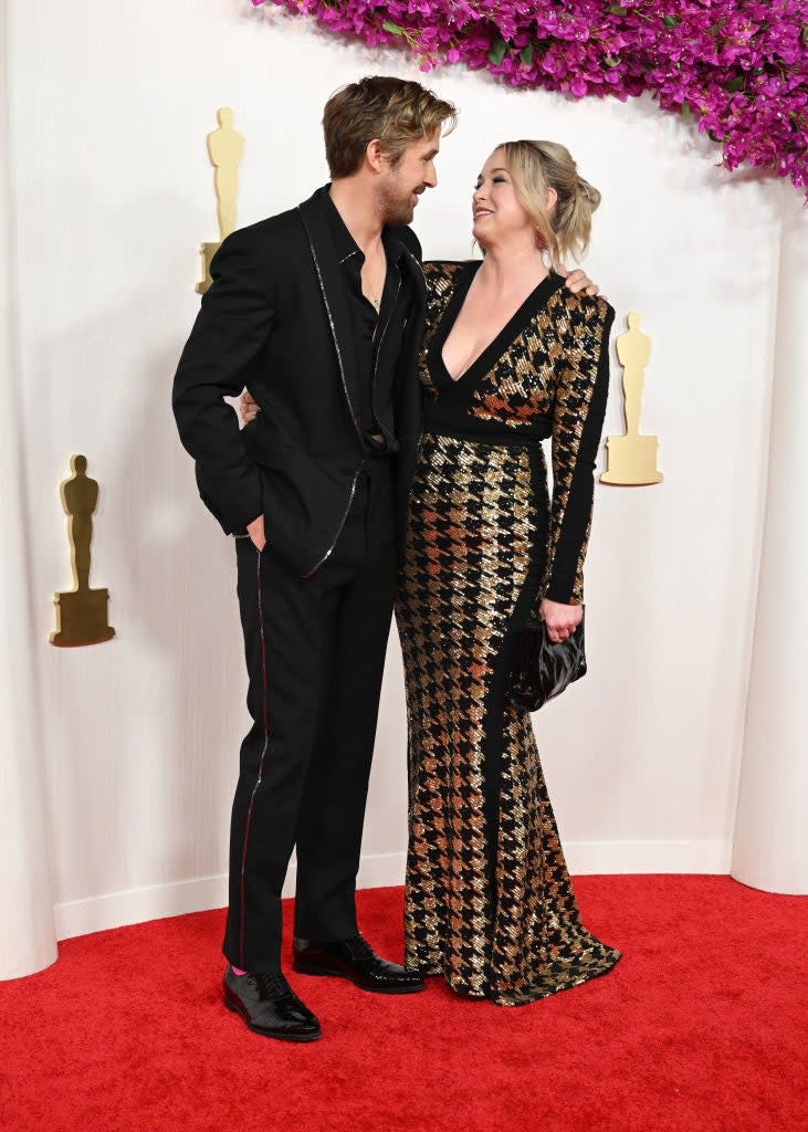 Two people on a red carpet, man in a black suit, woman in a patterned dress with a deep neckline. They are smiling at each other