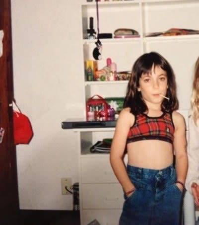 Young girl wearing jeans and a midriff-baring tank, with hands in jean pockets and a lollipop stick hanging out of their mouth