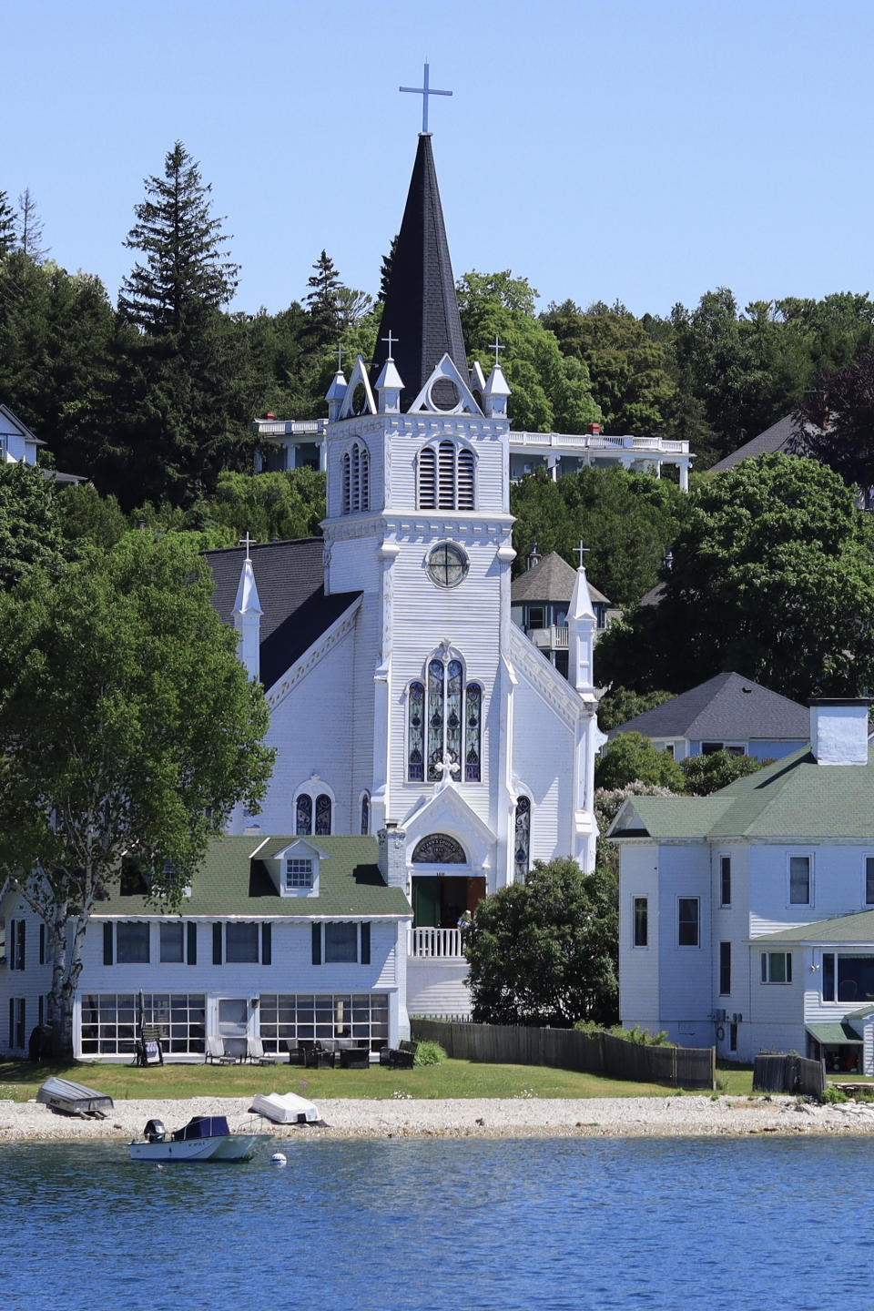 A white Gothic-style church with a tall spire is situated among trees and other buildings by a lakeside. A small boat is docked by the shore