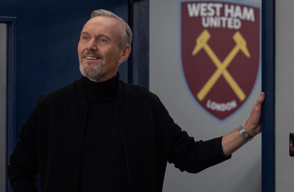 A man with grey hair and goatee wearing a black turtleneck and standing in a doorway with the West Ham United football logo on the wall behind him; still from "Ted Lasso"