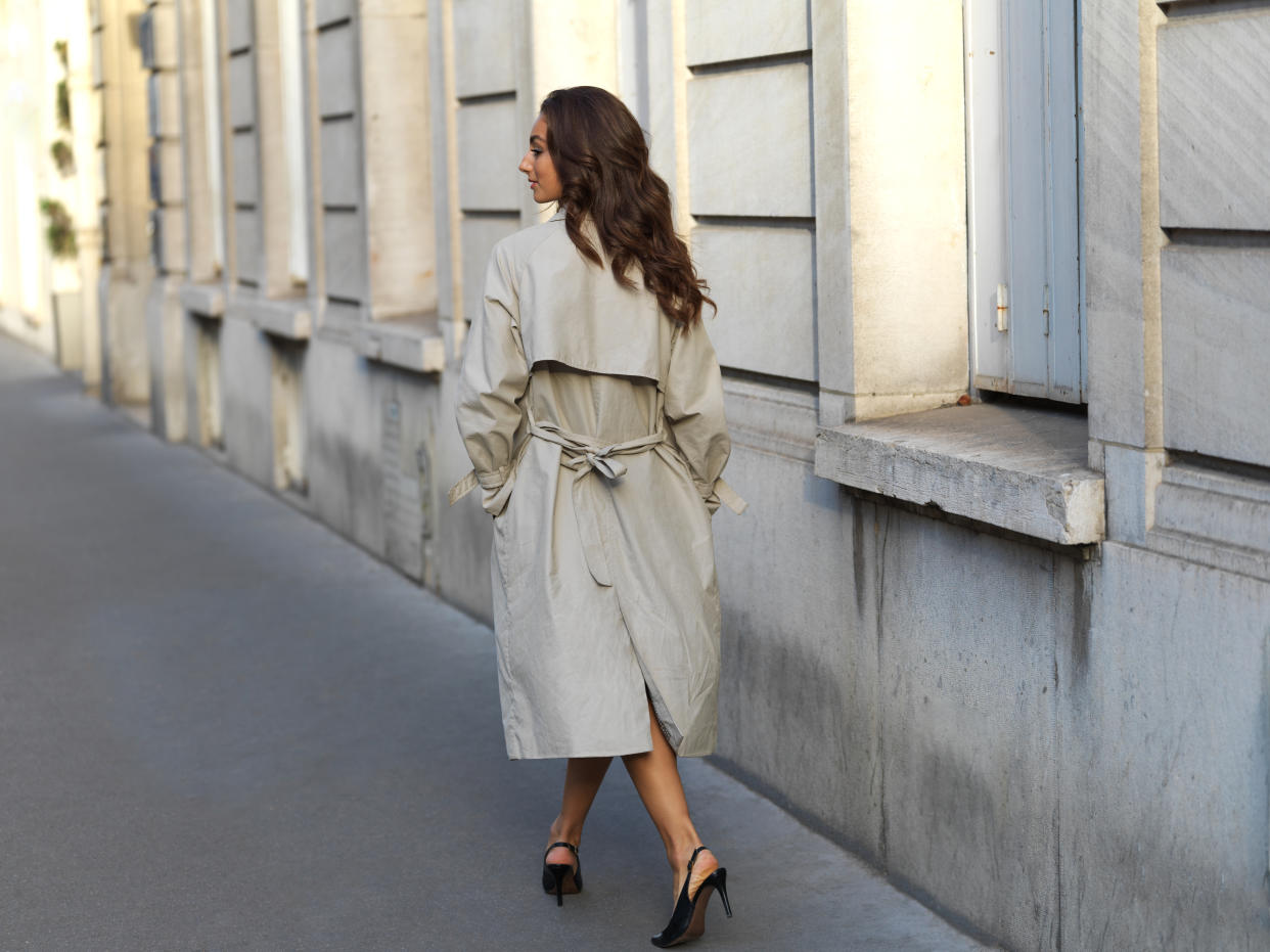 Stylish young woman wearing a trench coat, and high heels walking away from the camera on a sidewalk, street in Paris, France, on a beautiful bright sunny day