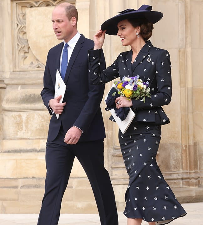 Kate Middleton holds on to her navy hat in a suit and matching skirt leaving Commonwealth Day at Westminster Abbey next to Prince William in a suit and royal blue tie