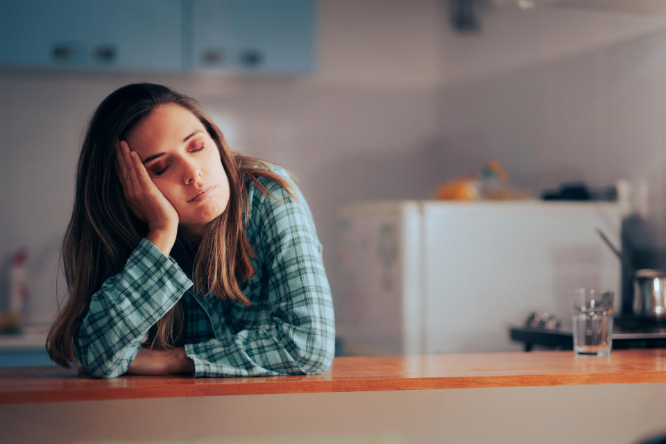 A woman in pajamas leans on a kitchen counter, resting her head on her hand with eyes closed, appearing tired