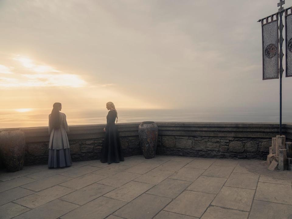 sonoya mizuno and emma d'arcy as mysaria and rhaenyra on house of the dragon, standing on a stone balcony overlooking a sunset. mysaria is wearing a blue dress, while rhaenyra is in black. they're facing each other and standing several feet apart